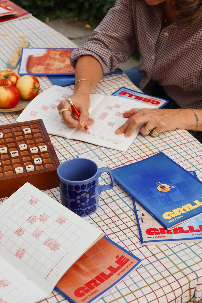 Femme complétant une grille de mots fléchés Grillé avec un stylo rouge, entourée de cahiers, d'une tasse bleue et de pommes
