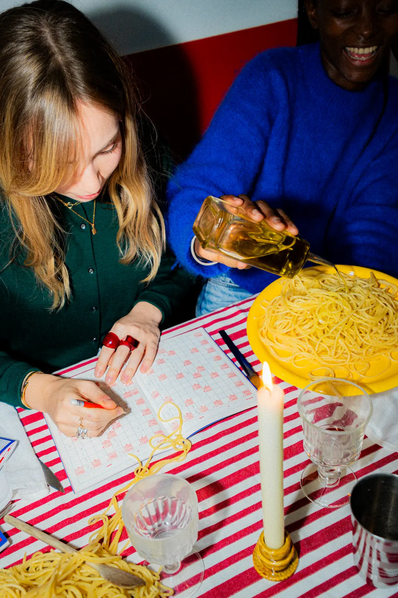 Femme remplissant un cahier de mots fléchés GRILLÉ pendant un dîner de pâtes, ambiance conviviale avec bougie et nappe rayée