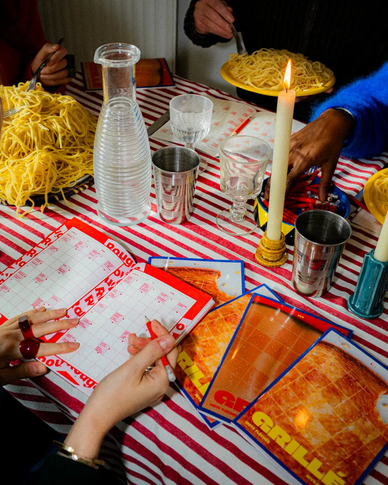 Cahier de mots fléchés GRILLÉ ouvert sur une table de dîner avec nappe rayée rouge et blanc, bougie allumée et plats de pâtes entre amis