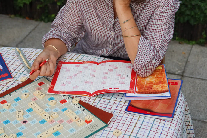 Personne complétant un cahier de mots fléchés Grillé avec stylo rouge sur table, parmi grilles de jeu