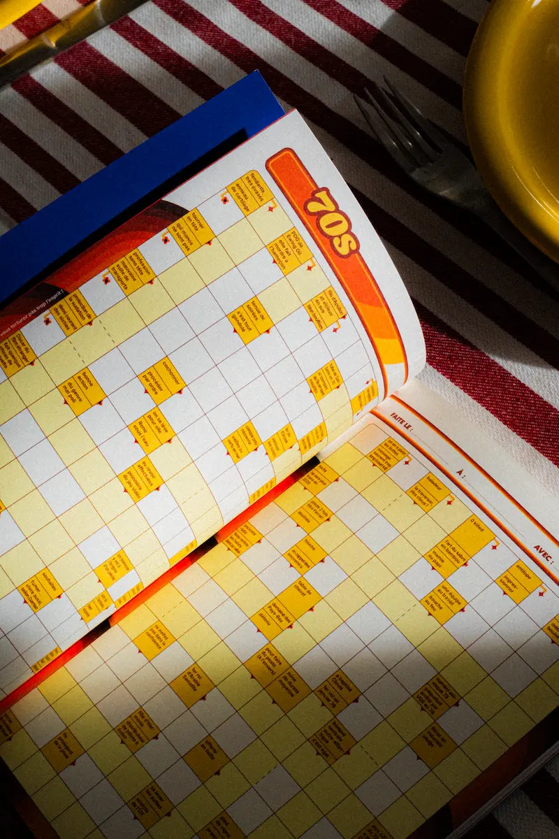 Cahier de mots fléchés GRILLÉ ouvert sur une grille thématique années 70, design rétro orange et jaune sur nappe rayée rouge et blanc