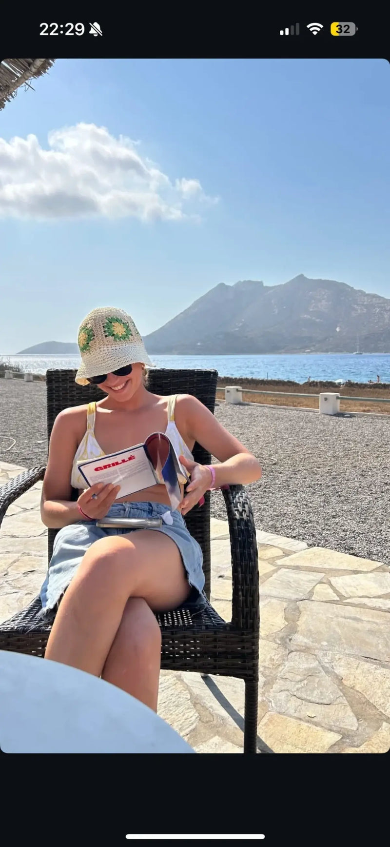 Femme souriante lisant un cahier de mots fléchés GRILLÉ face à la mer en Grèce